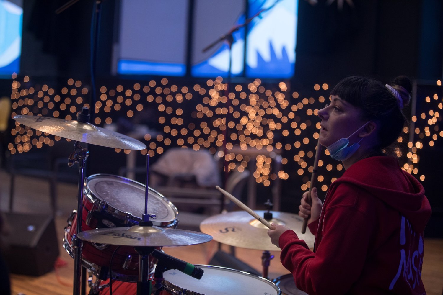 Rachel Parsons sits at a drum kit, ready to play, against a backdrop of twinkling fairy lights